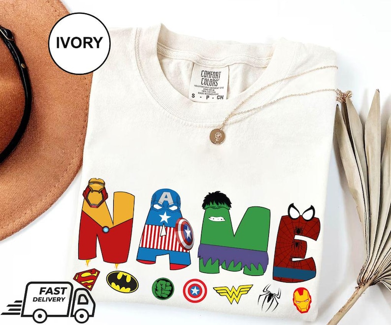 May include: Ivory-colored t-shirt with the word "NAME" in colorful superhero-themed letters. The shirt features various superhero logos and a gold necklace. The shirt is made of Comfort Colors material.
