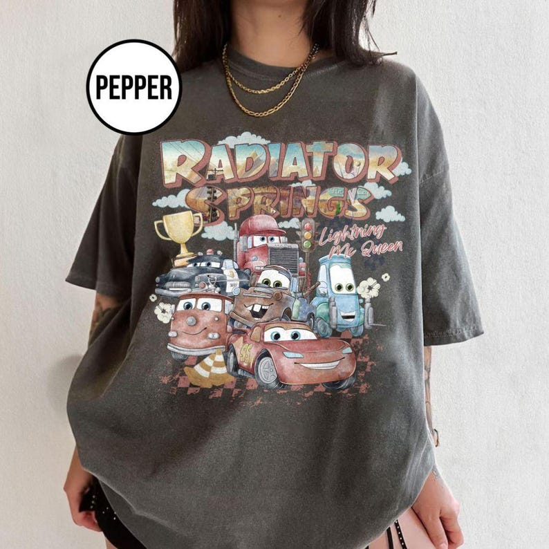 May include: Dark gray t-shirt featuring a colorful graphic of characters and the text "Radiator Springs" and "Lightning McQueen". The design includes a trophy and various cartoon cars. The shirt has short sleeves.