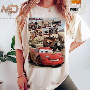 K&ouml;nnte beinhalten: Ein wei&szlig;es T-Shirt mit einer Grafik im Vintage-Stil von Lightning McQueen und Mater aus dem Disney Pixar Film Cars. Die Grafik lautet "Greetings from Radiator Springs".