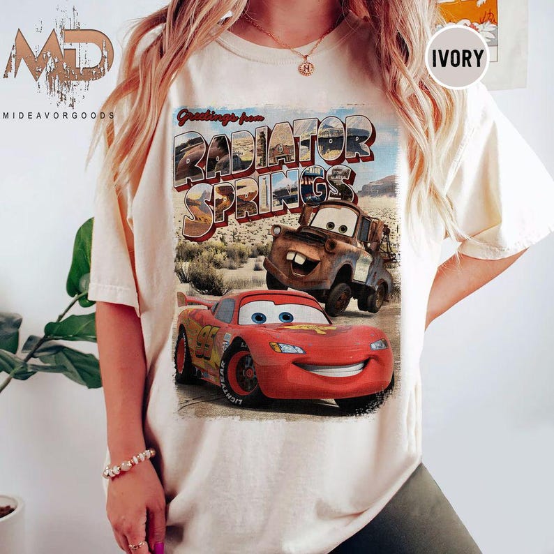 K&ouml;nnte beinhalten: Ein wei&szlig;es T-Shirt mit einer Grafik im Vintage-Stil von Lightning McQueen und Mater aus dem Disney Pixar Film Cars. Die Grafik lautet "Greetings from Radiator Springs".