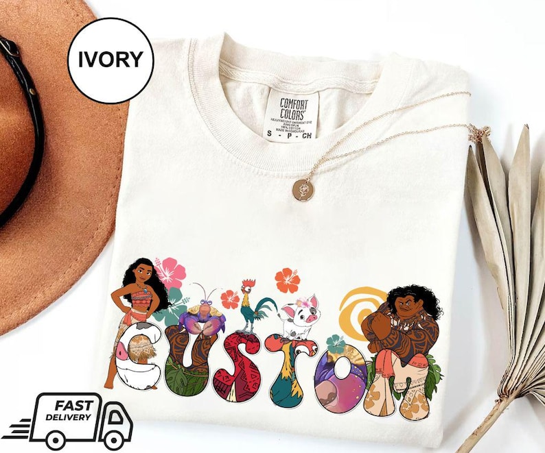 May include: Ivory t-shirt with a custom design featuring characters from the animated film Moana. The design includes Moana, Maui, Pua the pig, Hei Hei the rooster, and a stylized "CUSTOM" text.