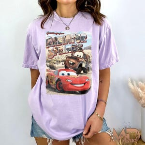 K&ouml;nnte beinhalten: Ein lavendelfarbenes T-Shirt mit einem Grafikdruck der Figuren Lightning McQueen und Mater aus dem Disney Pixar Film Cars. Die Grafik enth&auml;lt den Text "Greetings from Radiator Springs".