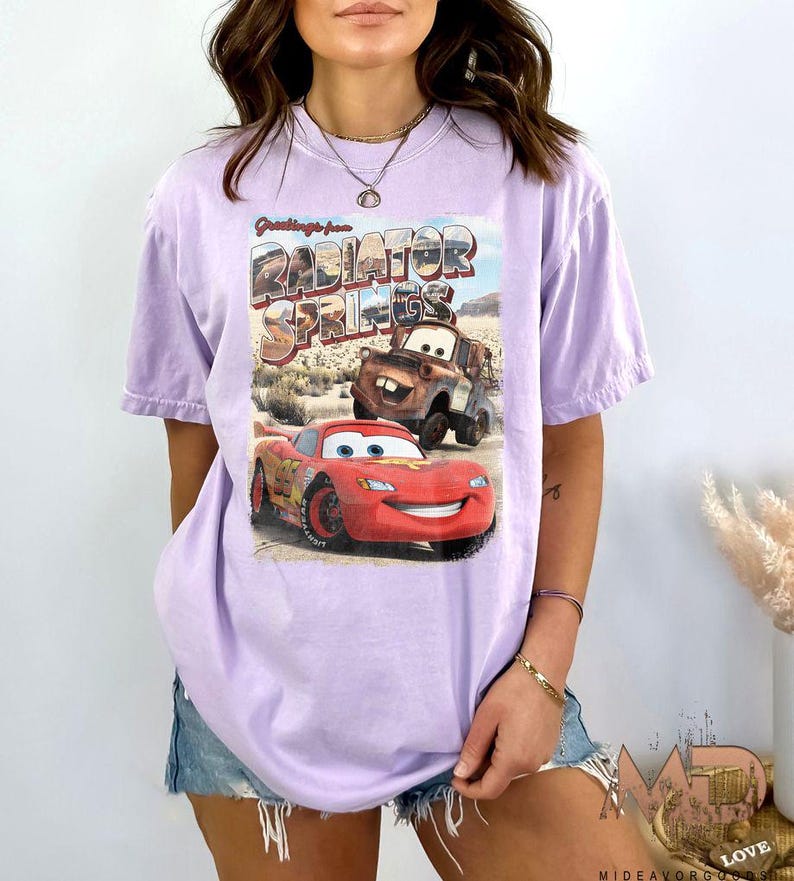 K&ouml;nnte beinhalten: Ein lavendelfarbenes T-Shirt mit einem Grafikdruck der Figuren Lightning McQueen und Mater aus dem Disney Pixar Film Cars. Die Grafik enth&auml;lt den Text "Greetings from Radiator Springs".