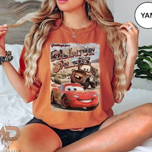 K&ouml;nnte beinhalten: Eine Frau tr&auml;gt ein orangefarbenes T-Shirt mit einem Aufdruck von Lightning McQueen und Mater aus dem Film Cars. Der Aufdruck lautet "Greetings from Radiator Springs".