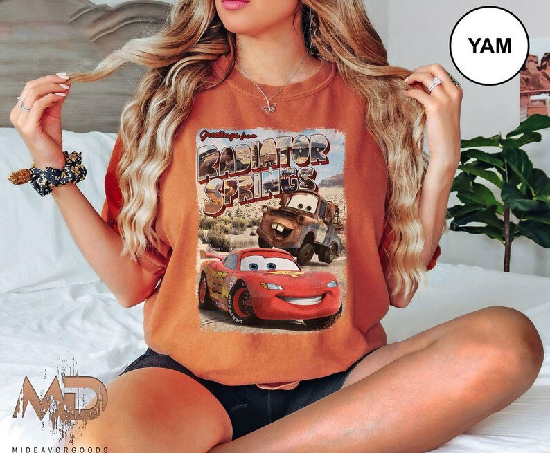 K&ouml;nnte beinhalten: Eine Frau tr&auml;gt ein orangefarbenes T-Shirt mit einem Aufdruck von Lightning McQueen und Mater aus dem Film Cars. Der Aufdruck lautet "Greetings from Radiator Springs".