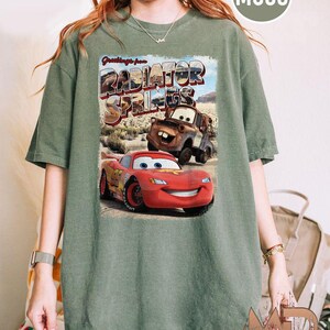 K&ouml;nnte beinhalten: Ein gr&uuml;nes T-Shirt mit einem Vintage-Postkarten-Design mit dem Text "Greetings from Radiator Springs". Die Postkarte zeigt ein rotes Rennwagen und einen rostigen blauen LKW.