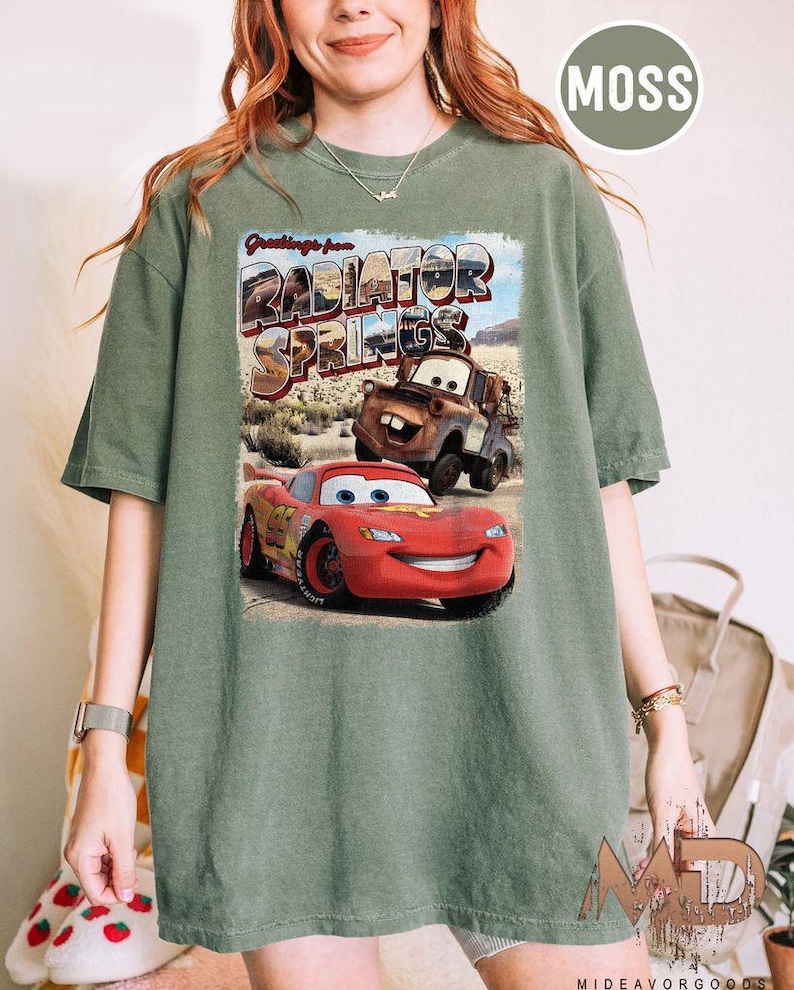 K&ouml;nnte beinhalten: Ein gr&uuml;nes T-Shirt mit einem Vintage-Postkarten-Design mit dem Text "Greetings from Radiator Springs". Die Postkarte zeigt ein rotes Rennwagen und einen rostigen blauen LKW.