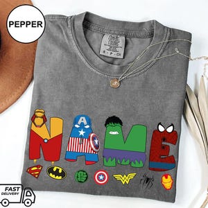 May include: A gray t-shirt with the word "NAME" in colorful superhero-themed letters. The letters are decorated with iconic symbols like Iron Man's mask, Captain America's shield, and the Hulk's face. A necklace and a hat are also visible.