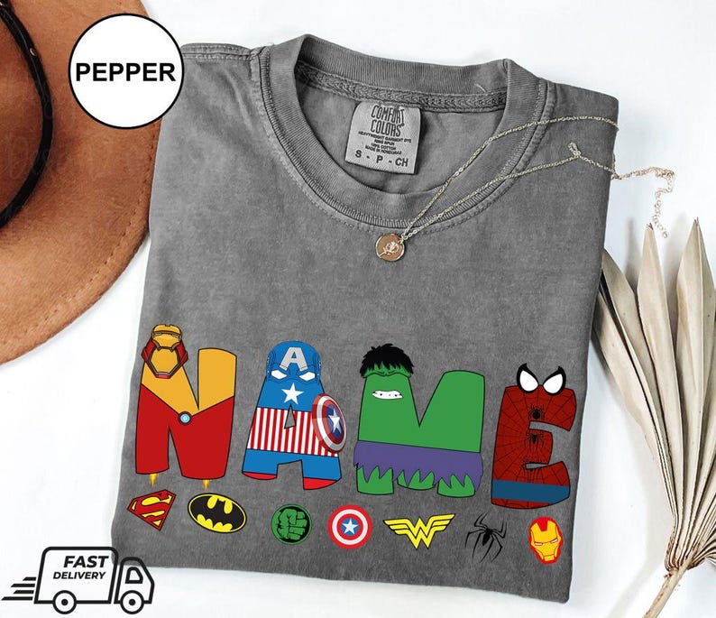 May include: A gray t-shirt with the word "NAME" in colorful superhero-themed letters. The letters are decorated with iconic symbols like Iron Man's mask, Captain America's shield, and the Hulk's face. A necklace and a hat are also visible.