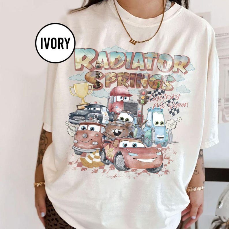 May include: Ivory-colored t-shirt featuring a graphic of characters from the movie Cars, including Lightning McQueen and Mater. The design includes the text "Radiator Springs" in a colorful, distressed font. A gold trophy and checkered flags are also visible.