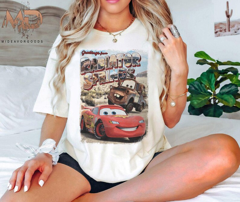 K&ouml;nnte beinhalten: Wei&szlig;es T-Shirt mit einem Vintage-Postkarten-Design mit dem Text "Greetings from Radiator Springs" und Bildern von Lightning McQueen und Mater aus dem Disney Pixar Cars Film.
