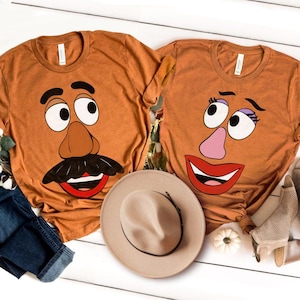 May include: Two orange t-shirts with cartoon faces, one with a mustache and the other with eyelashes and a pink nose. A tan hat and shoes are next to the shirts. The shirts are on a white wooden surface.