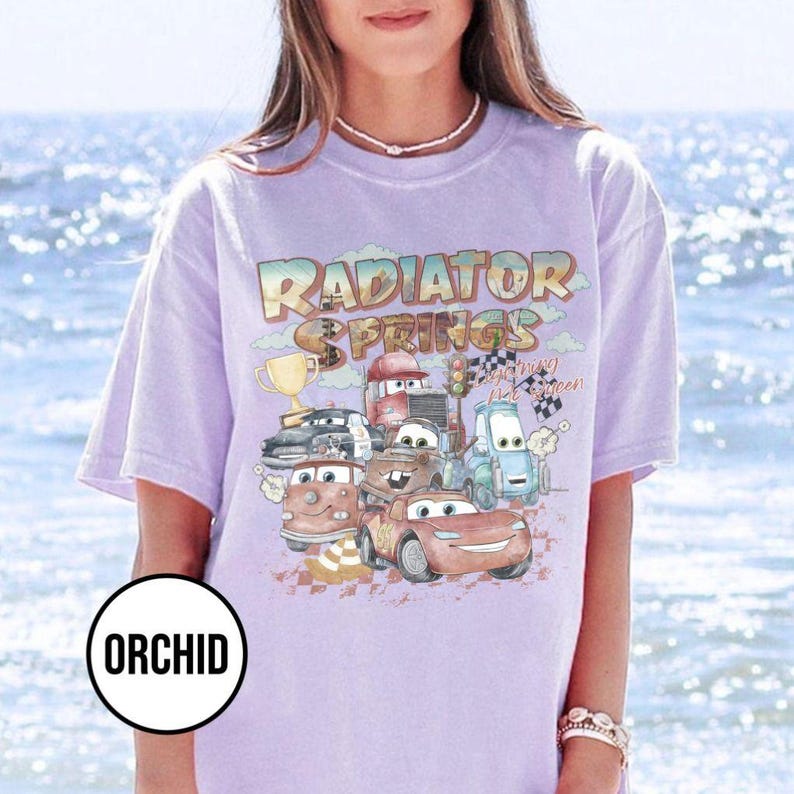 May include: A light purple t-shirt featuring a graphic of cartoon cars with the text "Radiator Springs". The design includes a trophy, checkered flags, and the words "Racing is my green". The shirt has short sleeves.