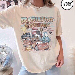 May include: Ivory-colored t-shirt featuring a colorful graphic of characters from the movie Cars, with the text "Radiator Springs". The design includes various animated cars, a trophy, and checkered flags, evoking a racing theme.