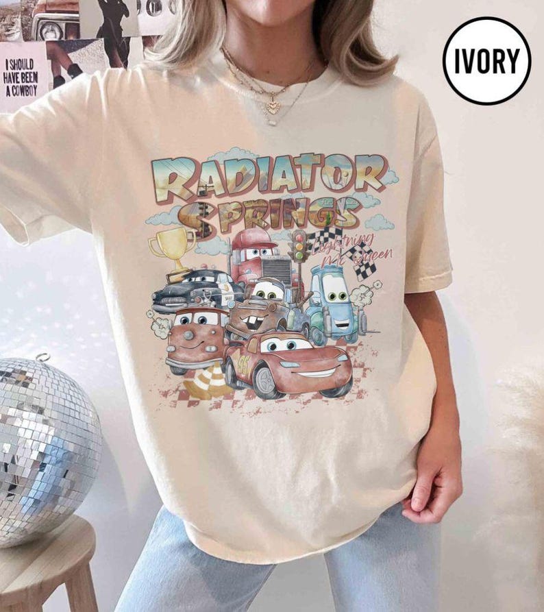 May include: Ivory-colored t-shirt featuring a colorful graphic of characters from the movie Cars, with the text "Radiator Springs". The design includes various animated cars, a trophy, and checkered flags, evoking a racing theme.