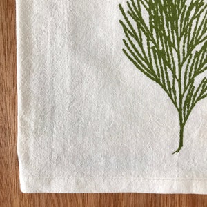 May include: A white kitchen towel with a green leafy design printed on it. The towel has a stitched hem.