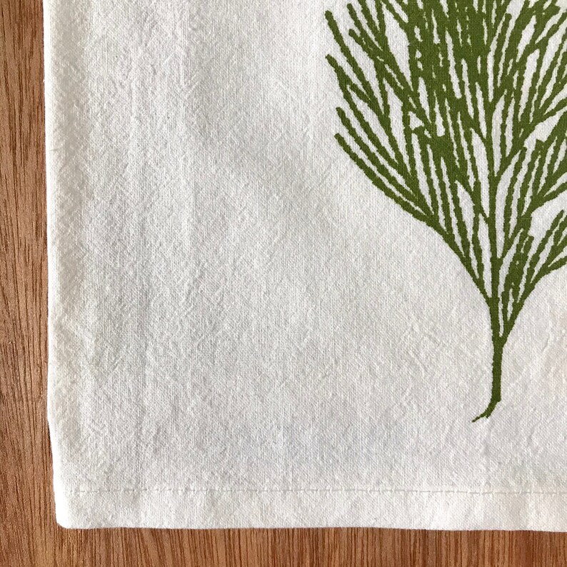 May include: A white kitchen towel with a green leafy design printed on it. The towel has a stitched hem.