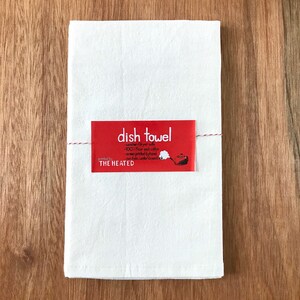 May include: White dish towel with a red label that says "dish towel" and includes details about the product, such as "washer/dryer safe", "100% flour sack cotton", "screen printed by hand", "non-fade, water-based", and "Printed by THE HEATED".