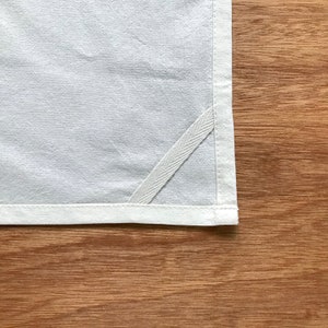 May include: A white kitchen towel with a white stitched border and a white ribbon sewn into the corner.
