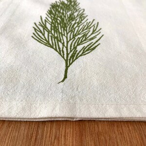 May include: A white kitchen towel with a green leafy design. The towel has a folded hem and is lying on a wooden surface.