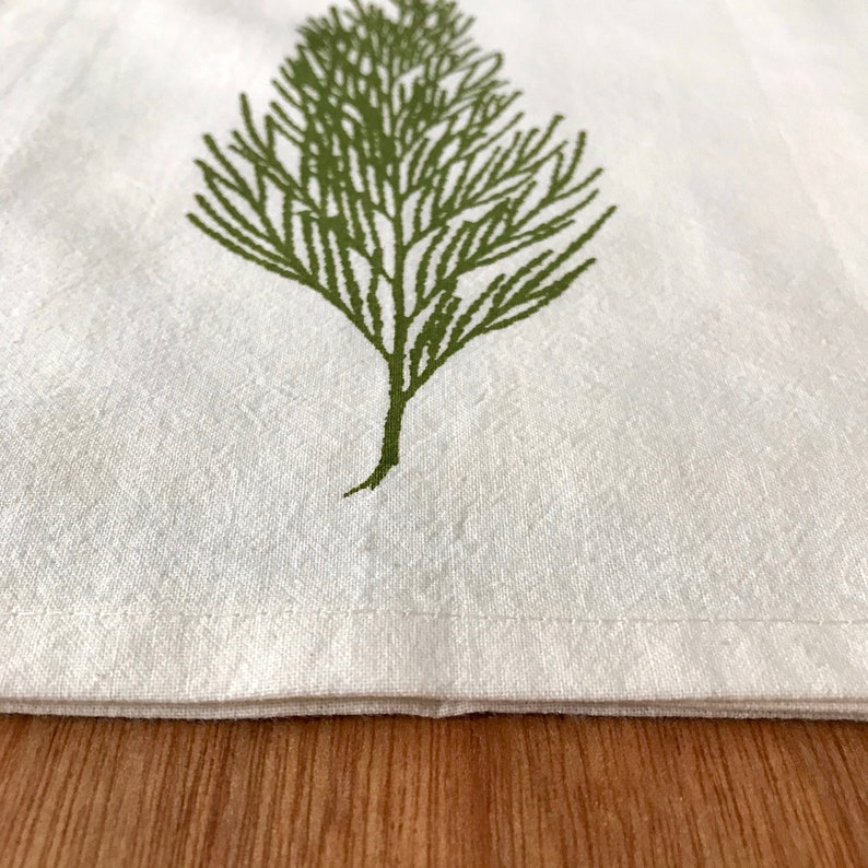 May include: A white kitchen towel with a green leafy design. The towel has a folded hem and is lying on a wooden surface.