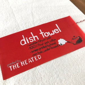 May include: Red dish towel with white text that reads "dish towel - washer/dryer safe - 100% flour sack cotton - screen printed by hand - non-toxic, water-based ink" and "printed by THE HEATED". A small illustration of a red teapot with steam coming out of the spout is in the upper right corner.