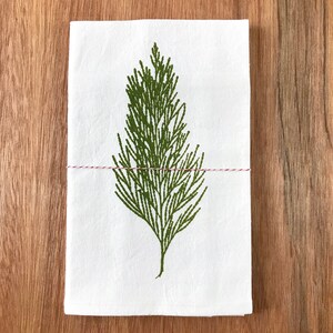 May include: A white kitchen towel with a green printed image of a pine branch. The towel is tied with a red and white string.