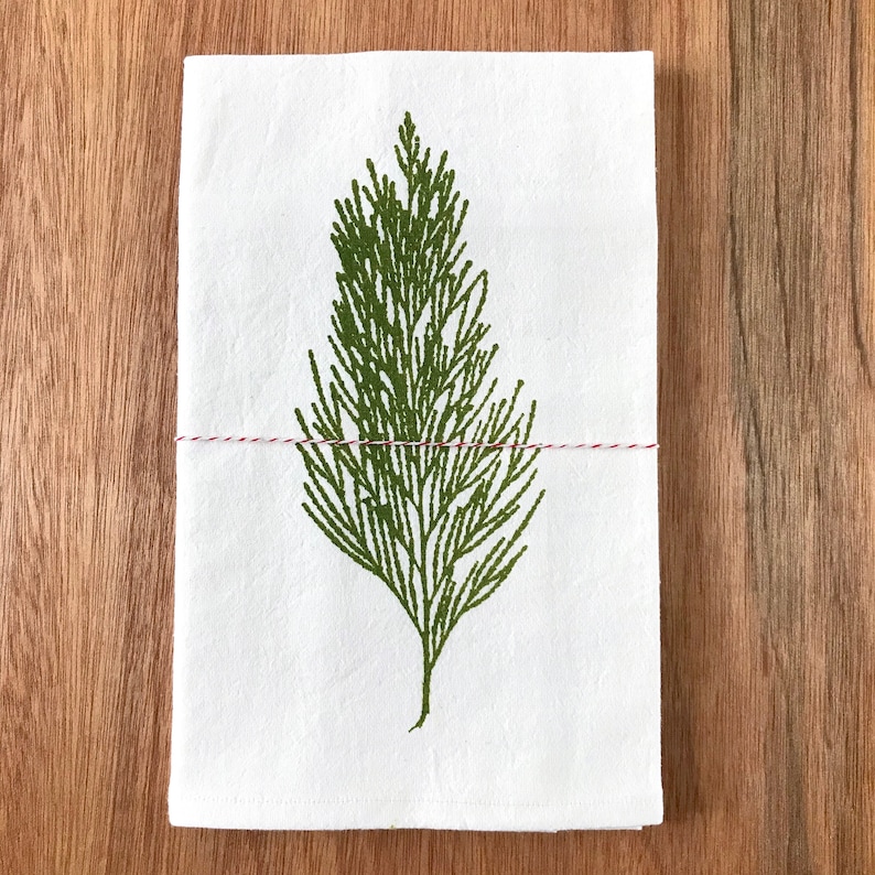 May include: A white kitchen towel with a green printed image of a pine branch. The towel is tied with a red and white string.