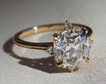 Könnte beinhalten: Ein goldener Ring mit einem großen, klaren, kissenförmigen Diamanten, der von kleineren Diamanten umgeben ist. Der Ring liegt auf einem weißen Stoffhintergrund.