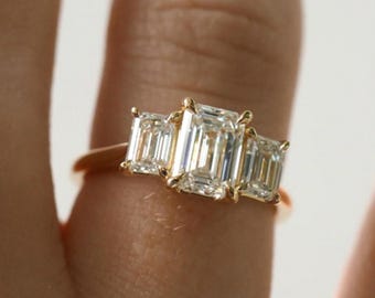 Puede incluir: Un anillo de oro con tres diamantes talla esmeralda. El anillo presenta un diseño clásico con una banda delicada. Los diamantes están engastados en fila, con la piedra central ligeramente más grande. Un símbolo de elegancia y sofisticación.