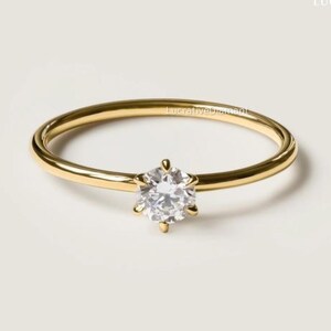 May include: A gold-colored ring with a single, clear, round-cut gemstone. The ring band is thin and smooth, and the gemstone is held in place by six prongs. The ring is set against a neutral background.