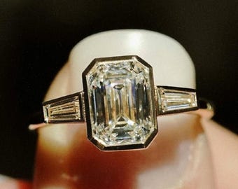 Puede incluir: Un anillo de compromiso de oro con un gran diamante de talla esmeralda y dos diamantes baguette más pequeños en la banda.