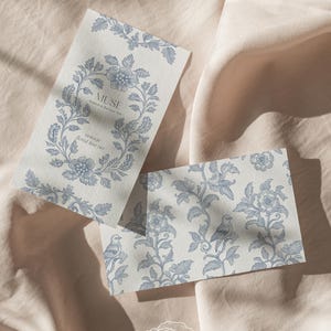 Op de afbeelding: Twee kaarten met een blauw en wit bloemenpatroon. De bovenste kaart heeft het woord "MUSE" in een cirkel met de tekst "Inspired in Baroque Style" eronder. De onderste kaart heeft een kleiner bloemenpatroon met vogels. De kaarten liggen op een lichtroze stoffen achtergrond.