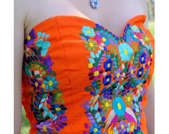 May include: A strapless orange top featuring vibrant, multi-colored floral embroidery in shades of blue, yellow, purple, and green. The garment is paired with a gold-tone chain necklace adorned with small white pearls, dark blue beads, and a blue and white pendant.
