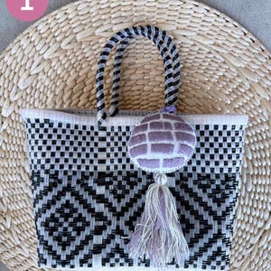May include: A woven tote bag with a black and white geometric pattern. The bag has a black and white twisted handle and a decorative purple tassel. The bag is sitting on a woven mat.