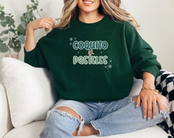 May include: Dark green sweatshirt with the text "COQUITO y PASTELES" in white and light green. The sweatshirt has a crew neck and long sleeves. The text is decorated with snowflakes.