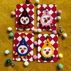 Puede incluir: Cuatro colgantes con forma de payaso de colores sobre tarjetas a cuadros rojos y blancos. Cada colgante tiene un esquema de color diferente: negro, rosa, azul y amarillo. Pequeños pompones y abalorios rodean las tarjetas.