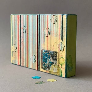 May include: A rectangular, handmade wooden box with a vintage aesthetic. The box features vertical stripes in various colors, including red, blue, and yellow. It has flower-shaped cutouts and a small, rectangular image of a woman. The sides are painted green.