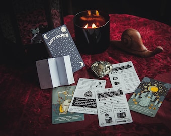 Puede incluir: Un juego de cartas de tarot con ilustraciones de una persona, una luna y un sol. Las cartas están dispuestas sobre una superficie de terciopelo rojo con un pequeño caracol de madera, una vela y una caja de papel de regalo. La caja de papel de regalo tiene un diseño de luna y sol y dice "Gift Paper" en la parte delantera.