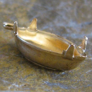 May include: A small, gold-colored boat-shaped jewelry finding. The finding has a rounded hull and raised edges with three vertical projections. The interior of the boat is hollow. The object is set against a mottled, neutral-toned surface.