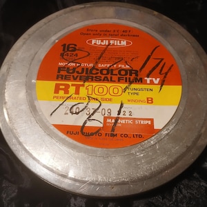 Puede incluir: Una bobina de película Fuji de 16 mm vintage con una etiqueta roja, amarilla y blanca. La etiqueta incluye texto como "FUJICOLOR REVERSAL FILM TV RT100" y marcas manuscritas. La bobina está alojada en un contenedor de metal plateado.
