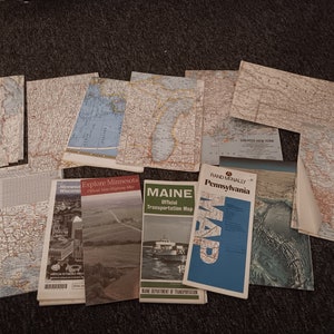 May include: A collection of vintage maps, including state highway maps and a Rand McNally Pennsylvania map. The maps feature detailed geographical information and are arranged on a dark surface. Some maps have text such as "Explore Minnesota" and "MAINE".