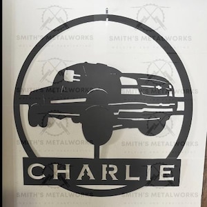 May include: Black metal wall art featuring a truck silhouette within a circular frame. The name "CHARLIE" is displayed below the truck. The words "SMITH'S METALWORKS" are repeated around the circle.