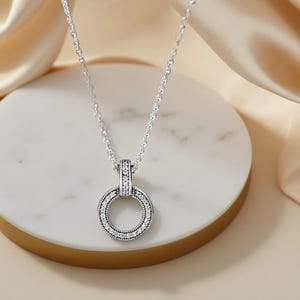 May include: A silver necklace featuring a circular pendant. The pendant is embellished with small, clear gemstones around the outer edge and a decorative section above. The necklace is displayed on a white and gold surface.
