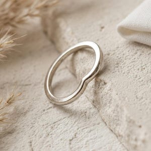 May include: A polished silver-toned ring featuring a smooth, continuous band with a subtle V-shaped chevron curve at the top. The metal surface is reflective and uniform, showcasing a simple, curved design that maintains a consistent thickness throughout.