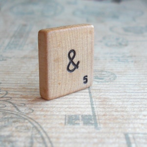 May include: A wooden Scrabble tile featuring the ampersand symbol (&) and the number 5 in black ink. The tile has a light brown color and is standing upright against a patterned background. This item is suitable for crafts and decorative purposes.