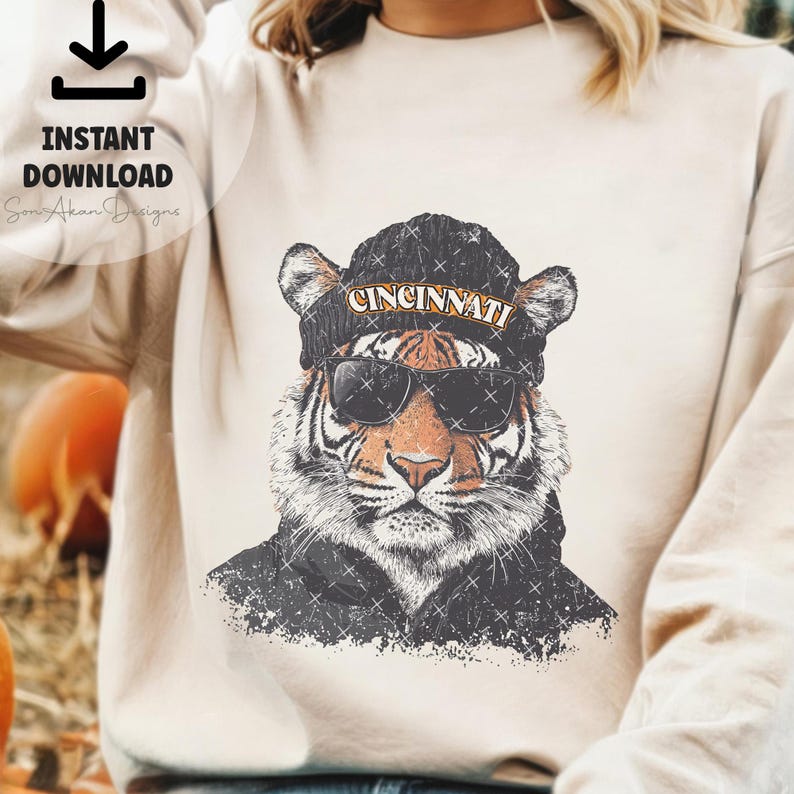 May include: A white sweatshirt featuring a graphic of a tiger wearing a black beanie with the word "CINCINNATI" on it. The tiger is sporting sunglasses and has a distressed, vintage aesthetic.