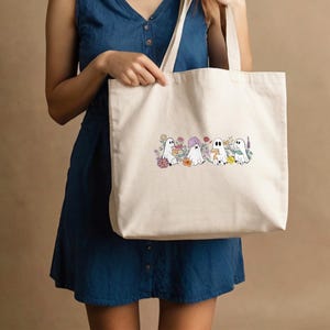 May include: A white canvas tote bag with a design of four ghosts holding books and surrounded by flowers. The ghosts are white with black eyes and mouths. The flowers are in various colors, including pink, purple, yellow, and orange.