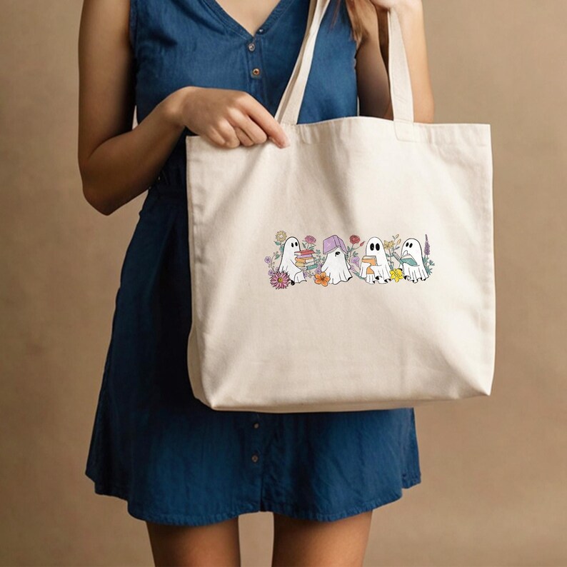 May include: A white canvas tote bag with a design of four ghosts holding books and surrounded by flowers. The ghosts are white with black eyes and mouths. The flowers are in various colors, including pink, purple, yellow, and orange.