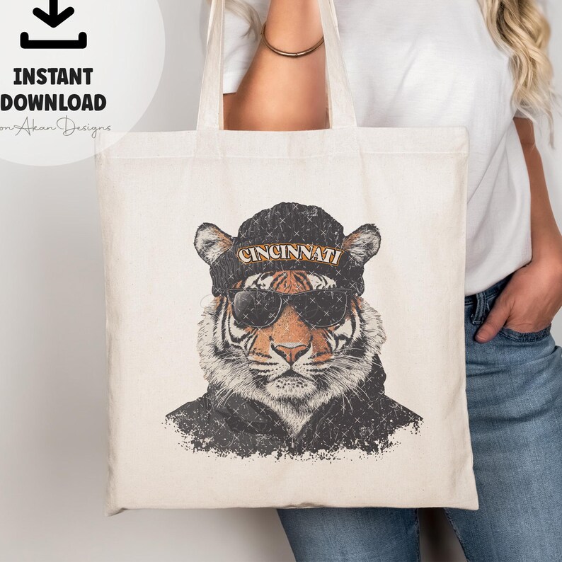 May include: A white canvas tote bag featuring a graphic of a tiger wearing a black beanie with the word "CINCINNATI" on it. The tiger is sporting sunglasses and has a distressed, vintage aesthetic.